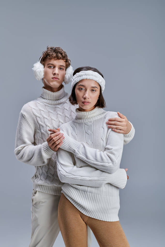 man in de winter oorkappen omarmen trendy aziatische vrouw in trui op grijs, mode lookbook concept - Foto, afbeelding