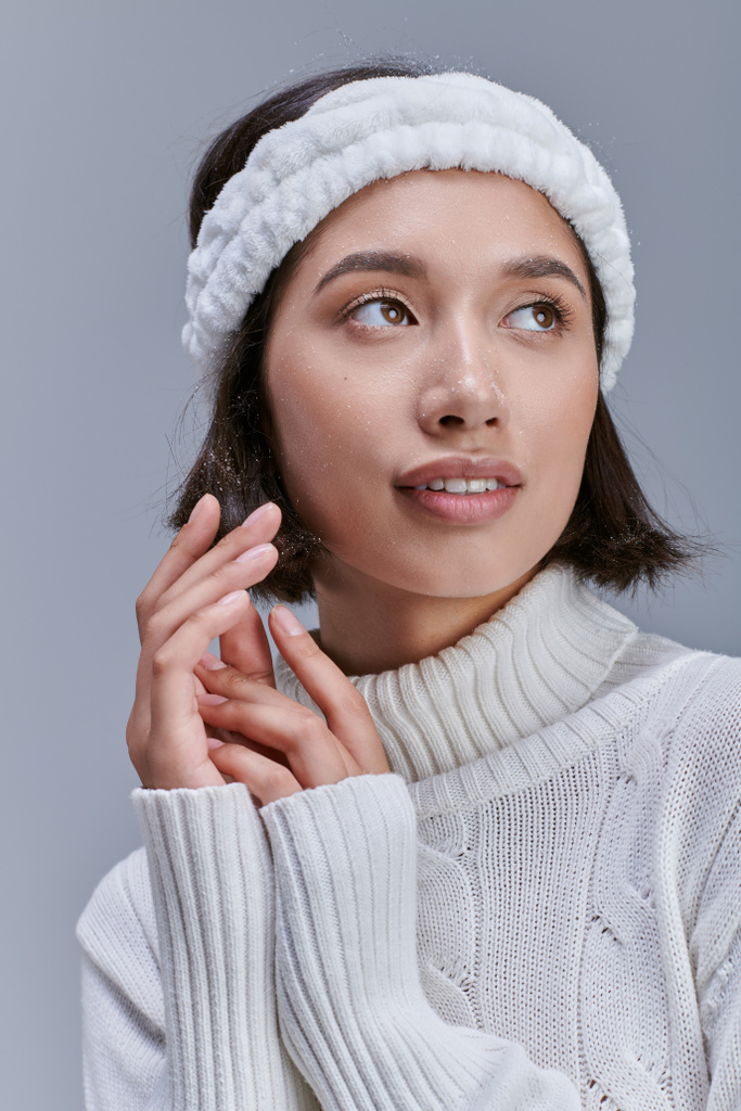 mujer asiática soñadora en suéter de punto y diadema suave mirando hacia otro lado en gris, tendencia de invierno moderno - Foto, Imagen