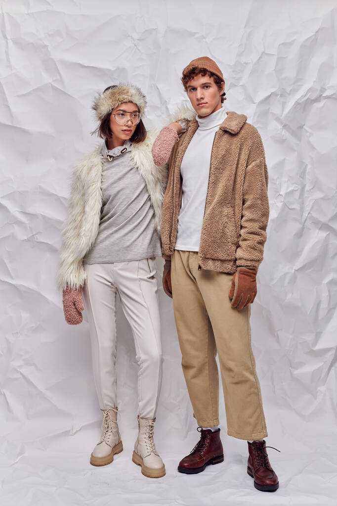 полная длина межрасовой пары в теплой одежде, стоящей на белом текстурированном фоне, зимний стиль - Фото, изображение