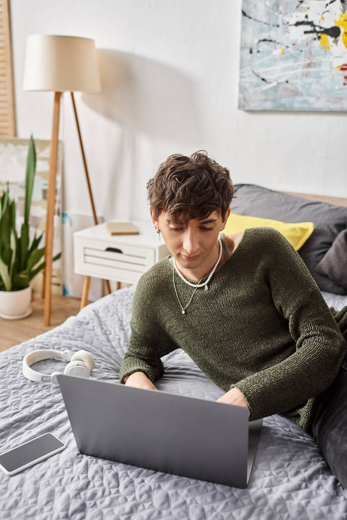 молодий кучерявий трансгендерний блогер використовує ноутбук і сидить на ліжку біля смартфона та навушників - Фото, зображення