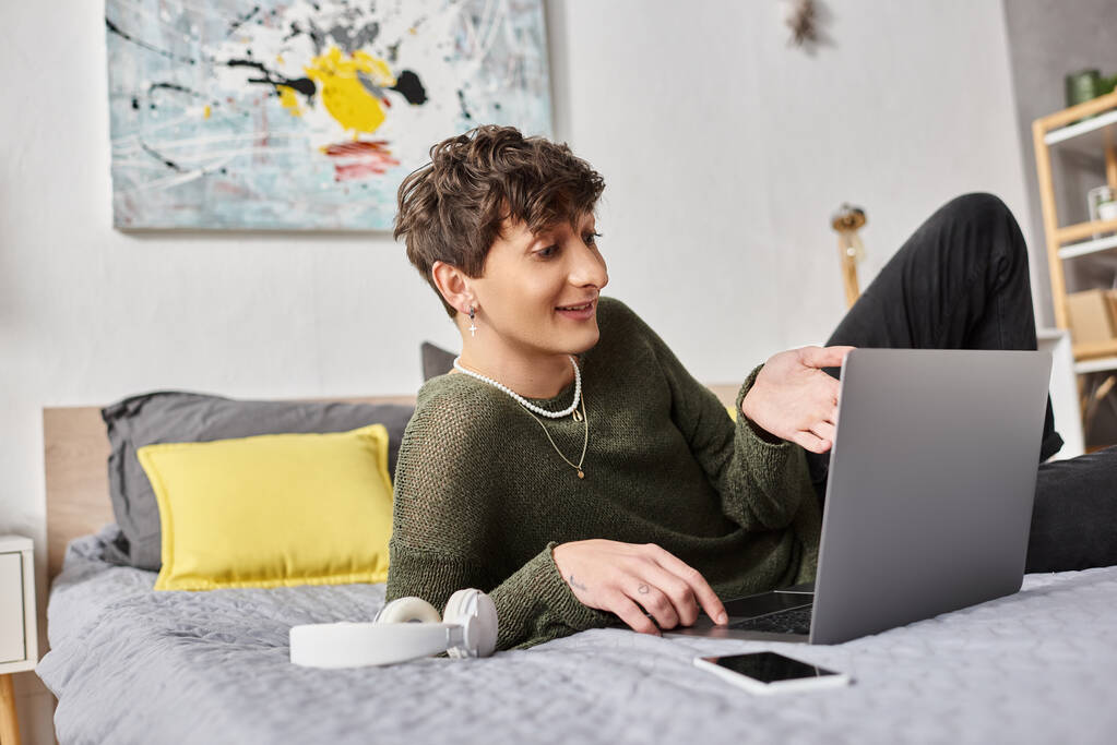 onnellinen kihara transsukupuolinen bloggaaja kannettavan tietokoneen avulla ja istuu sängyssä älypuhelimen ja kuulokkeiden lähellä - Valokuva, kuva