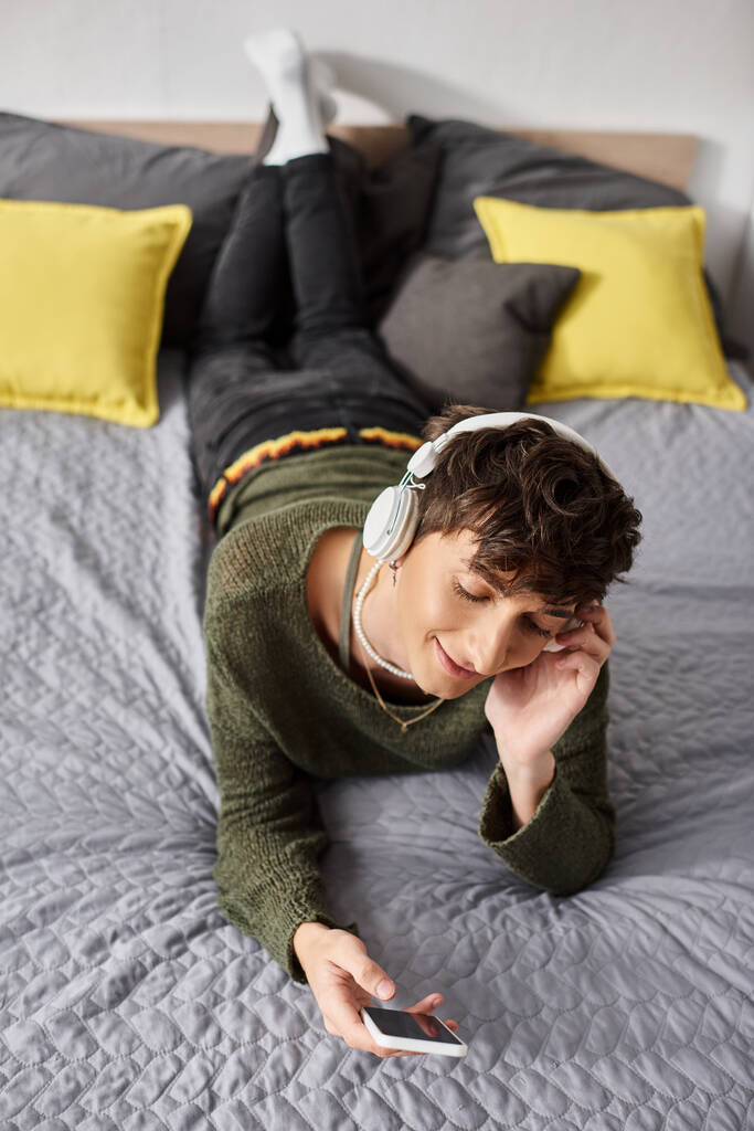 щаслива кучерява трансгендерна людина в бездротових навушниках, що тримає смартфон у спальні, слухати музику - Фото, зображення