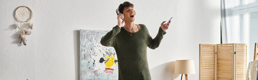 szczęśliwy kręcone transseksualista osoba w bezprzewodowych słuchawek słuchanie muzyki i trzymając smartfona, baner - Zdjęcie, obraz
