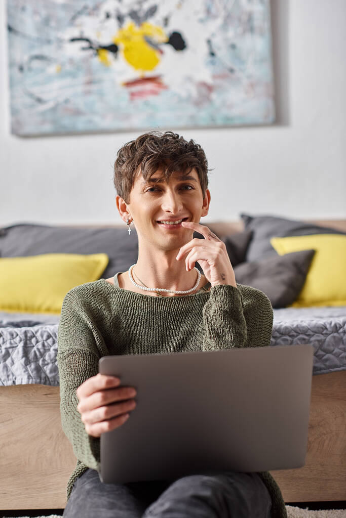 tevreden en krullend transgender influencer met behulp van laptop en zitten naast bed, sociale media - Foto, afbeelding
