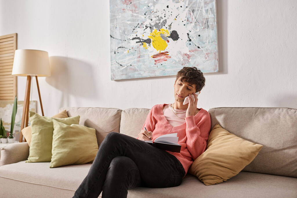 blogger transgénero en jersey rosa hablando en smartphone y escribiendo en notebook, sentado en sofá - Foto, imagen