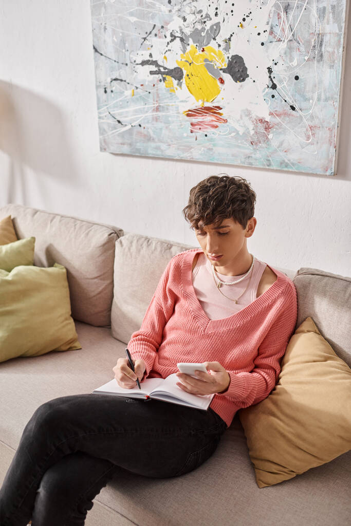 transsukupuolinen bloggaaja vaaleanpunaisessa puserossa pitäen älypuhelinta ja kirjoittaen muistikirjaan, istuen sohvalla - Valokuva, kuva