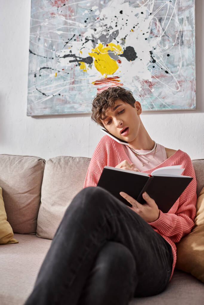 Transgender-Bloggerin im rosafarbenen Pullover, die auf dem Sofa sitzt und mit dem Smartphone spricht und in ein Notizbuch schreibt - Foto, Bild