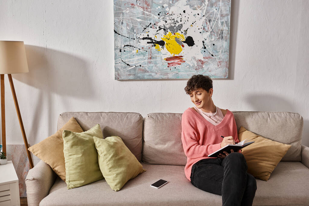 bloguero transgénero feliz en suéter rosa escrito en cuaderno y mirando el teléfono inteligente en el sofá - Foto, Imagen