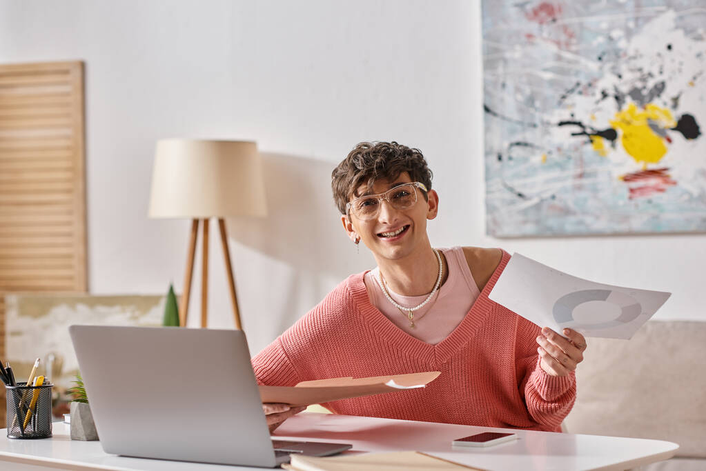gelukkig androgyne blogger in roze trui en bril werken op afstand in de buurt van apparaten op het bureau - Foto, afbeelding