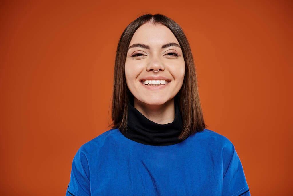gelukkig jong vrouw met doorboorde neus kijken naar camera en glimlachen op oranje achtergrond, blauwe jas - Foto, afbeelding