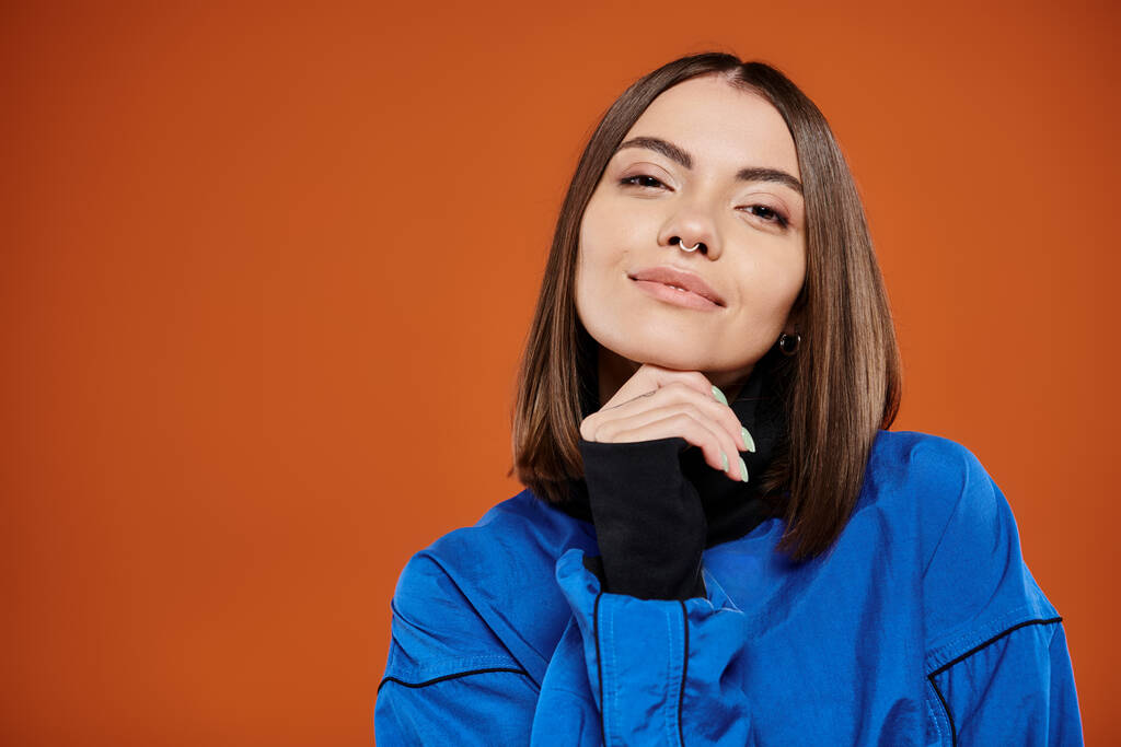 mujer pensativa con la nariz perforada mirando a la cámara mientras piensa en fondo naranja, chaqueta azul - Foto, imagen
