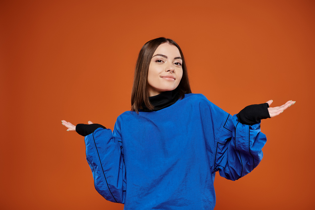 confused woman with pierced nose looking at camera while standing on orange backdrop, blue jacket - Photo, Image