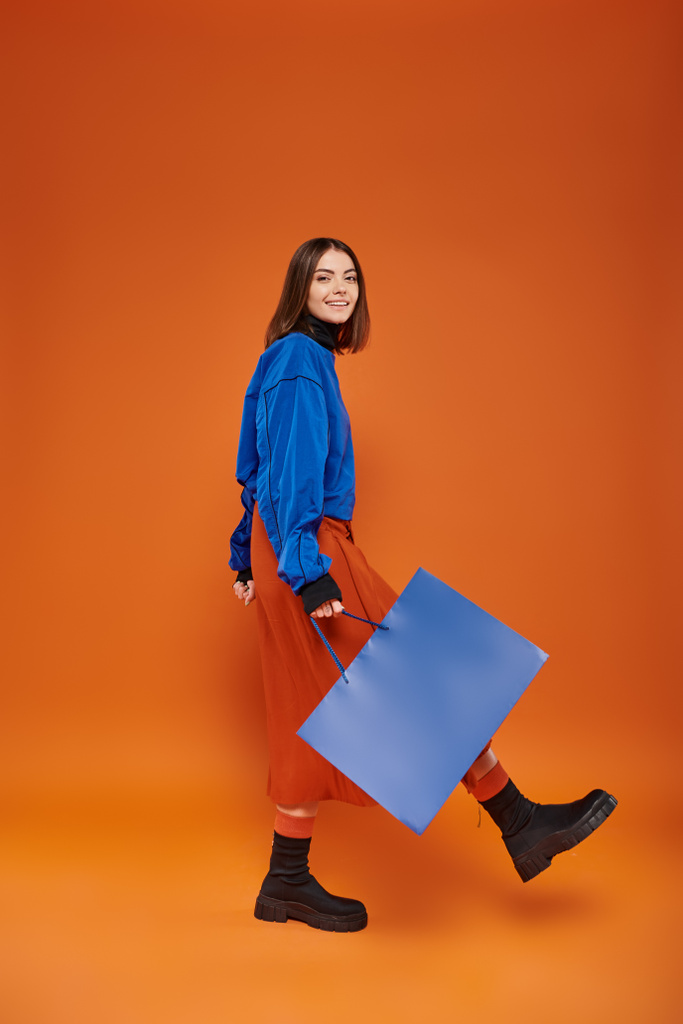 Sonbahar kıyafetleri içinde mutlu bir kadın turuncu arka planda alışveriş çantasıyla yürüyor. Kara Cuma konsepti. - Fotoğraf, Görsel