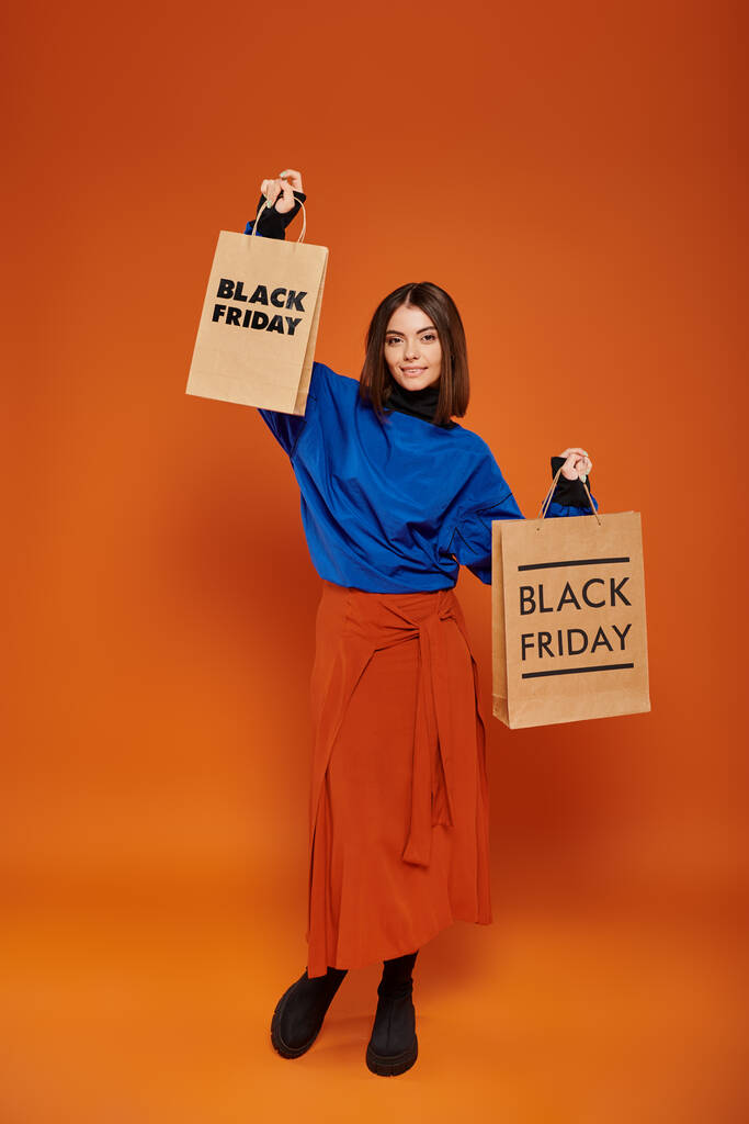 longitud completa, mujer bonita en traje de otoño sosteniendo bolsas de compras sobre fondo naranja, viernes negro - Foto, imagen