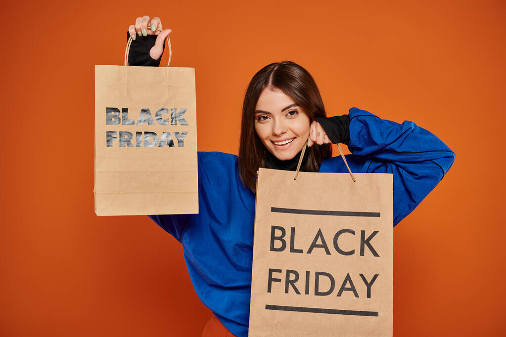 mujer alegre sosteniendo bolsas de compras con letras de viernes negro sobre fondo naranja, temporada de ventas - Foto, imagen