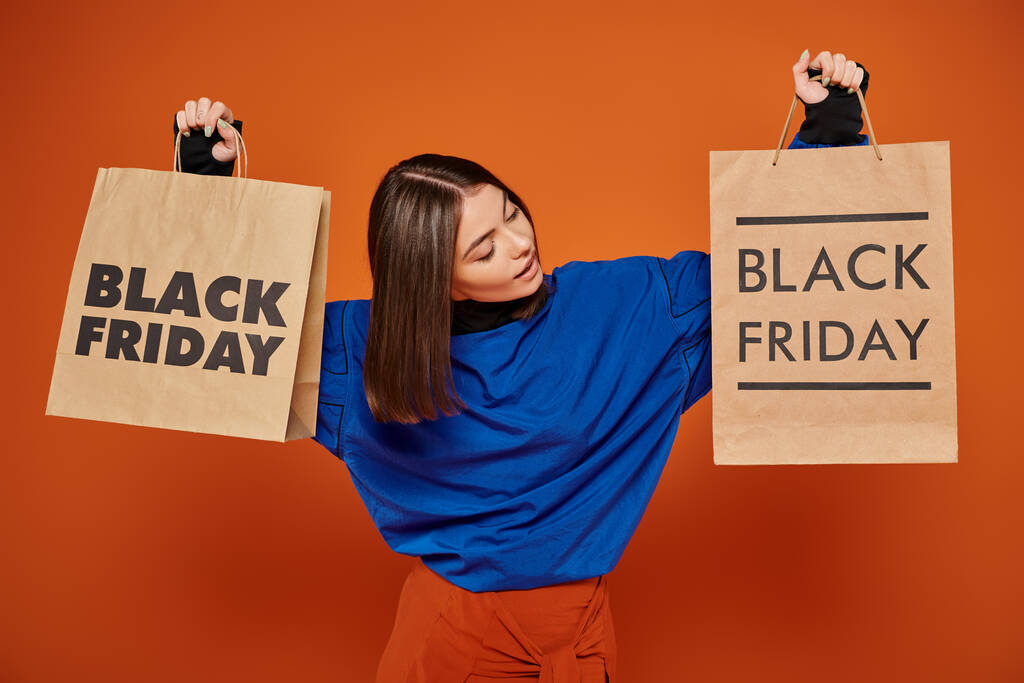 брюнетка женщина держит сумки с черными пятничными буквами на оранжевом фоне, сезон продаж - Фото, изображение