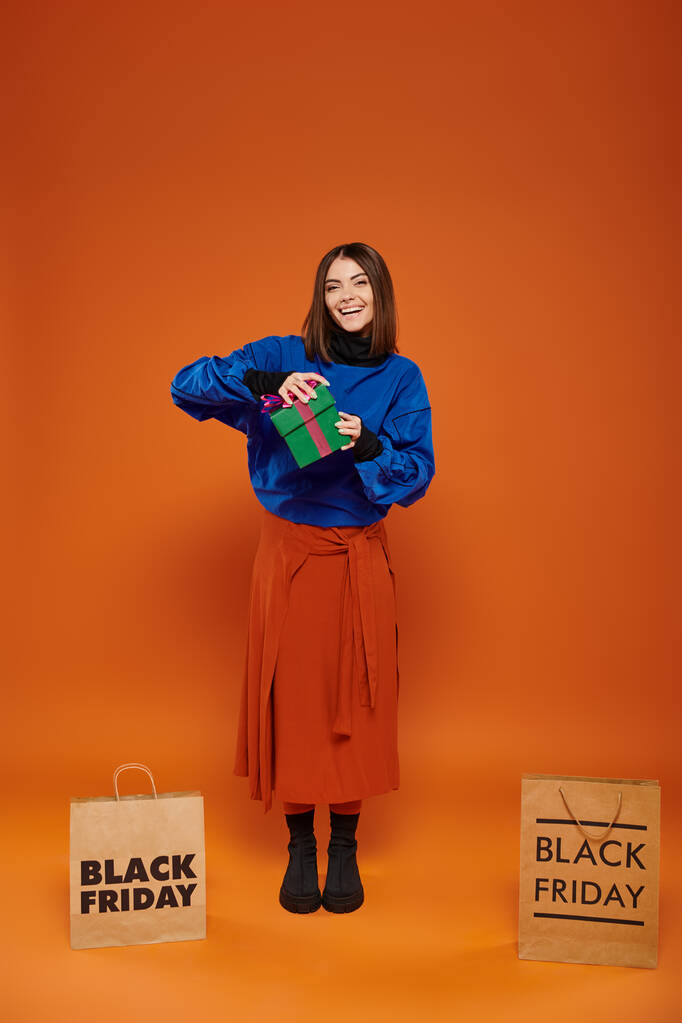 gelukkig vrouw opening cadeau in de buurt van boodschappentassen met zwarte vrijdag letters op oranje achtergrond - Foto, afbeelding