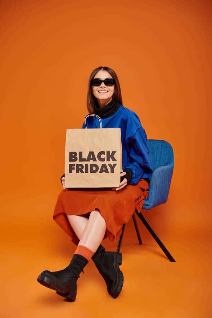 wesoła kobieta w stylowych okularach przeciwsłonecznych trzyma czarny piątkowy worek na zakupy i siedzi na fotelu - Zdjęcie, obraz