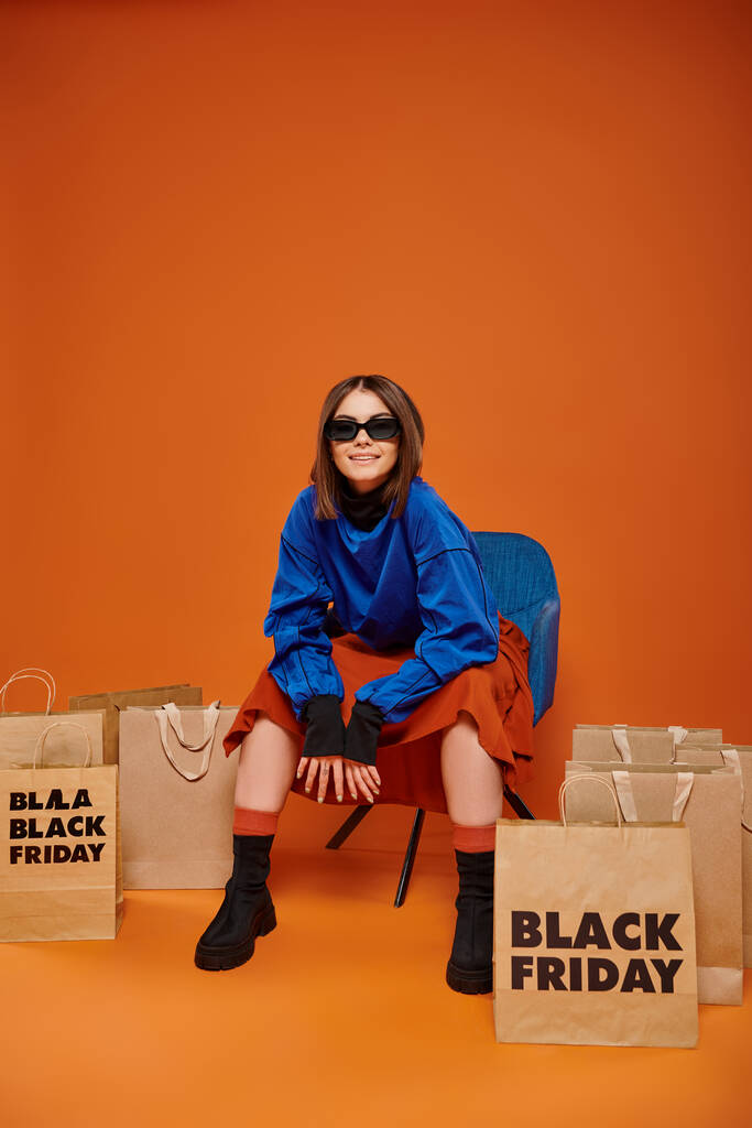 オレンジのブラックフライデーのショッピングバッグの近くのアームチェアに座っているスタイリッシュなサングラスの肯定的な女性 - 写真・画像