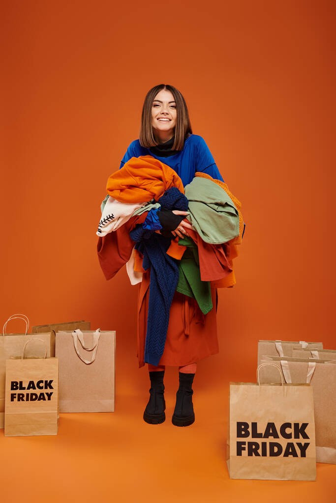 mujer alegre con cabello moreno sosteniendo ropa otoñal vibrante cerca de bolsas de compras de viernes negro - Foto, imagen