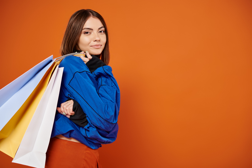 mujer positiva en ropa otoñal con estilo sosteniendo bolsas de compras el viernes negro, fondo naranja - Foto, imagen