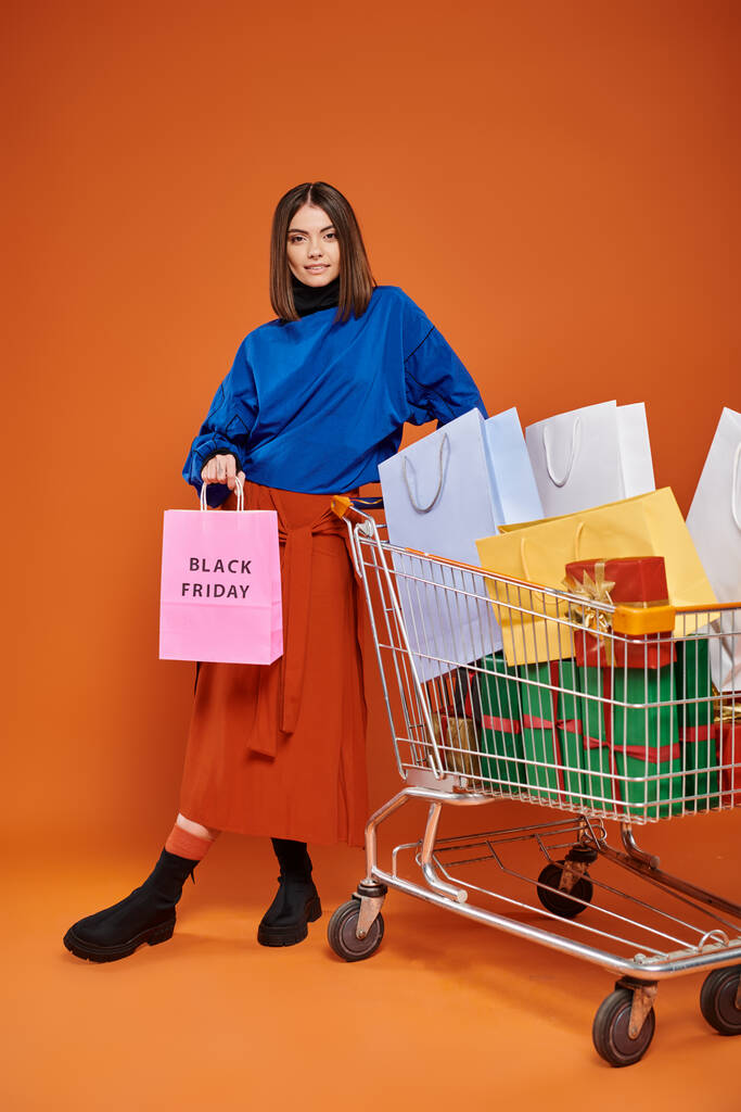 pretty woman standing with cart full of shopping bags with black friday letters on orange, wave hand - Photo, Image