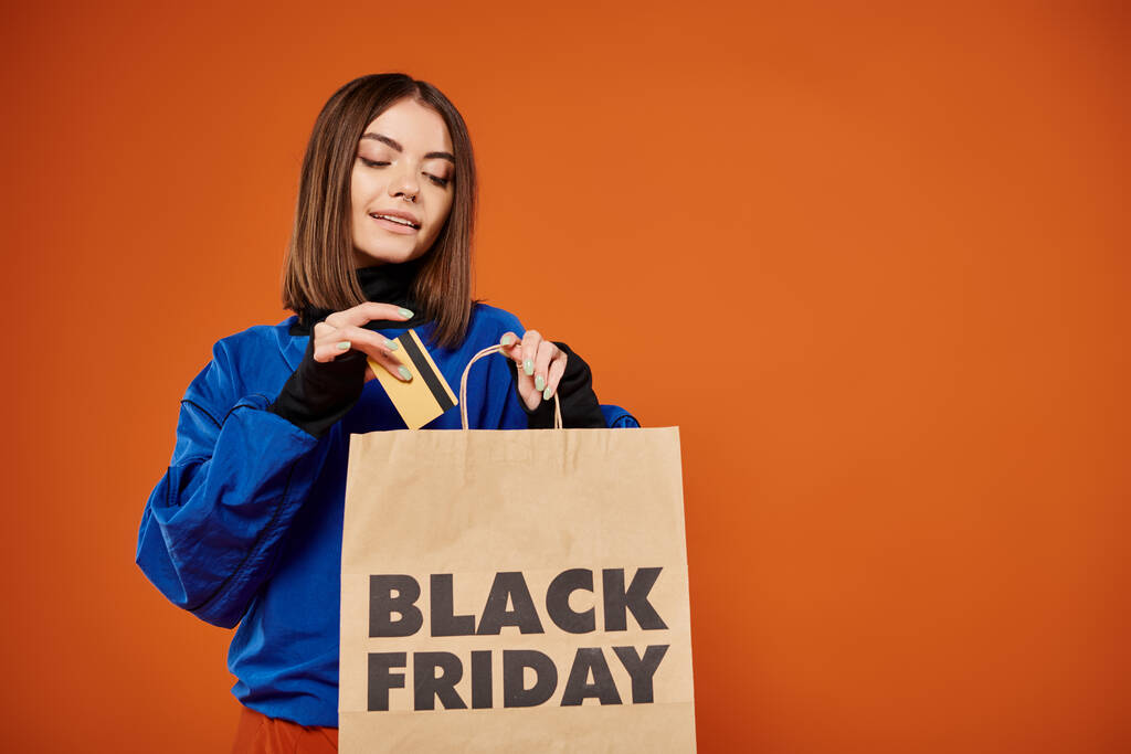 bella donna mettendo carta di credito in shopping bag con nero venerdì lettere su sfondo arancione - Foto, immagini