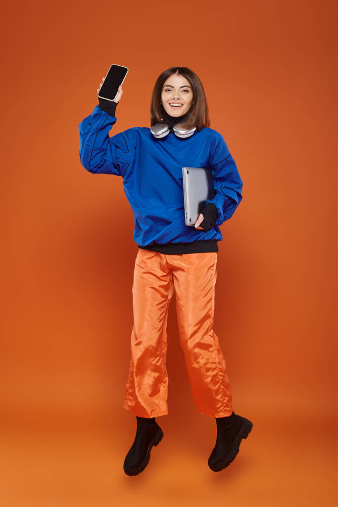 femme gaie en tenue d'automne tendance sauter avec des gadgets sur orange, concept cyber lundi - Photo, image