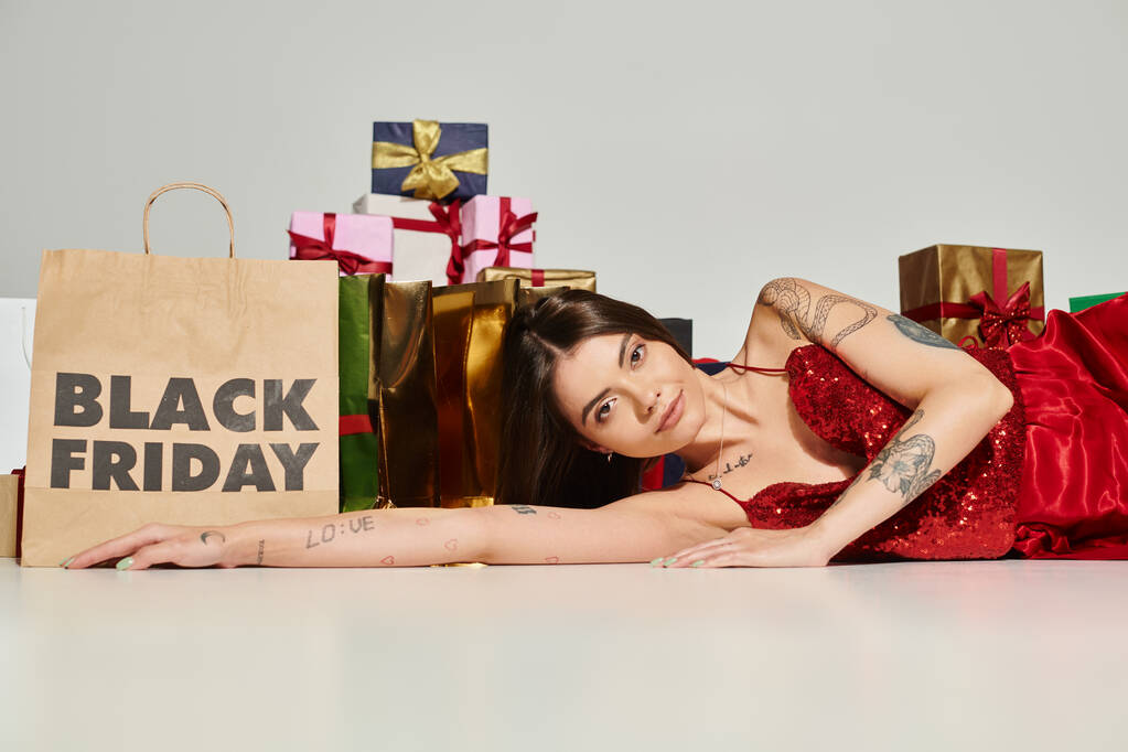 заманчивая молодая женщина позирует на полу возле подарков и сумки для покупок на фоне ecru, черная пятница - Фото, изображение
