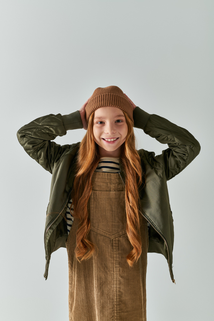 положительная девушка с длинными волосами носить зимнюю шляпу и стоя в платье с курткой на сером фоне - Фото, изображение