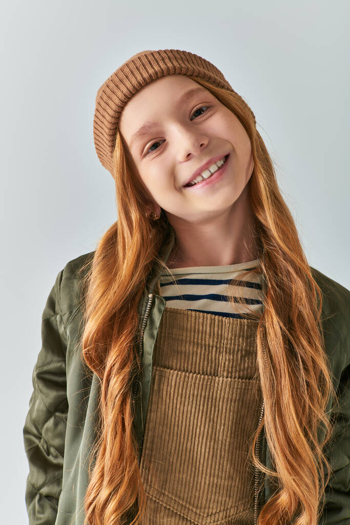 χειμωνιάτικη μόδα, χαρούμενο κορίτσι με πλεκτό καπέλο και εξωτερικά ενδύματα κοιτάζοντας την κάμερα σε γκρι φόντο - Φωτογραφία, εικόνα
