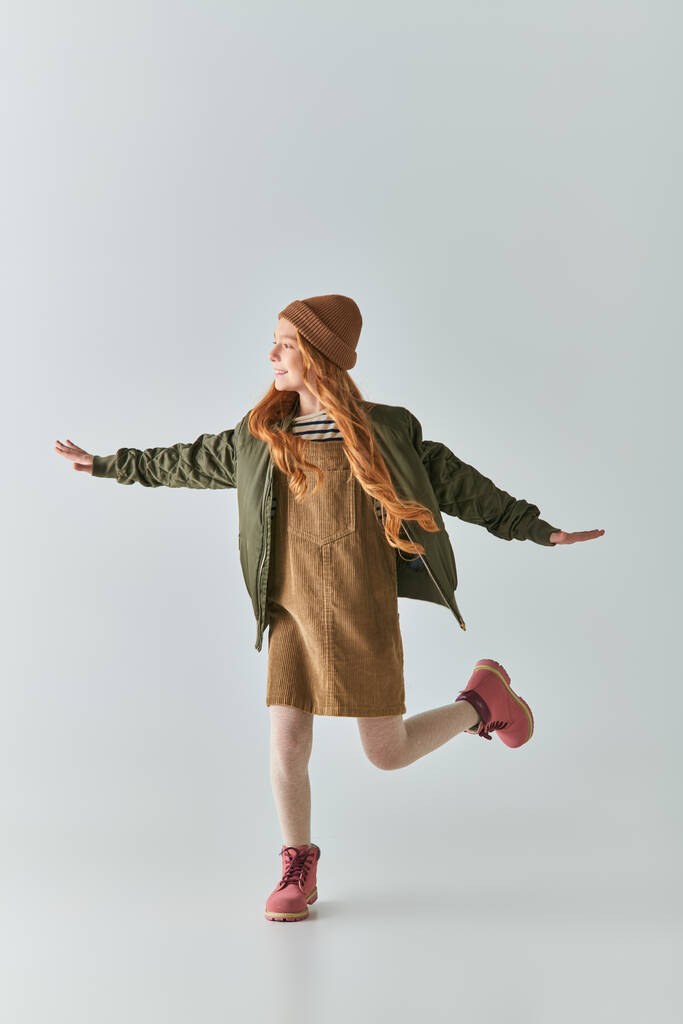 comprimento total da menina animado em roupa de inverno e chapéu de malha correndo com as mãos estendidas em cinza - Foto, Imagem