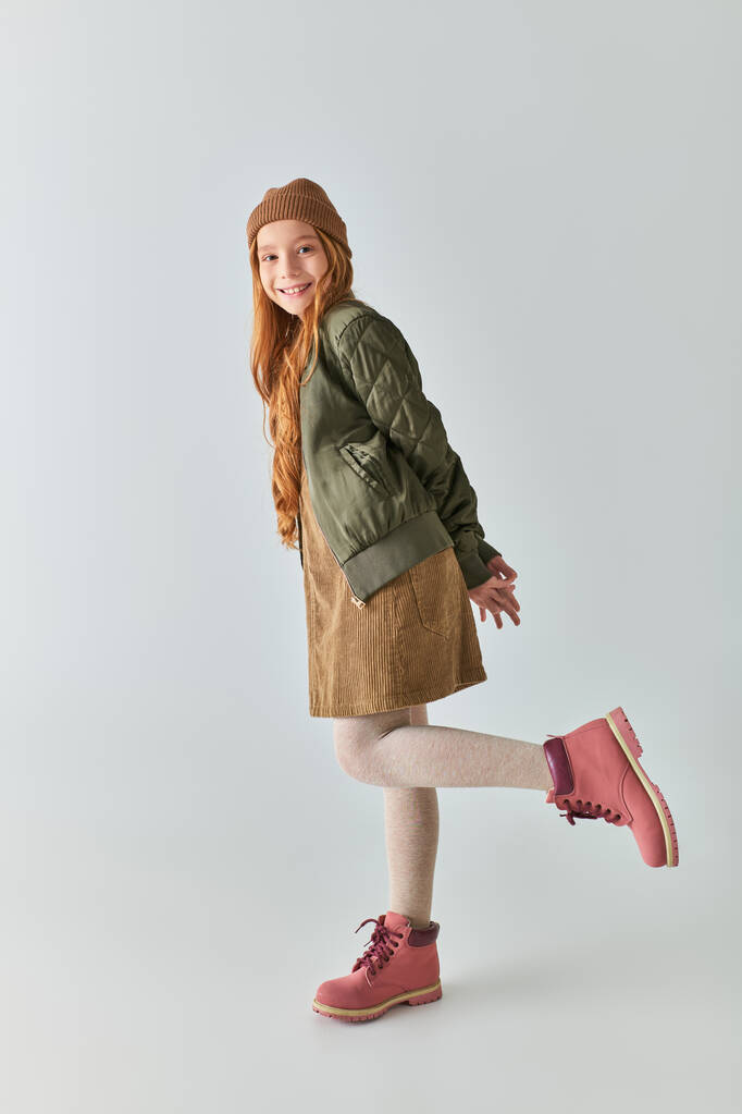 longitud completa de niña preadolescente alegre en traje de invierno con botas y sombrero mirando a la cámara en gris - Foto, Imagen