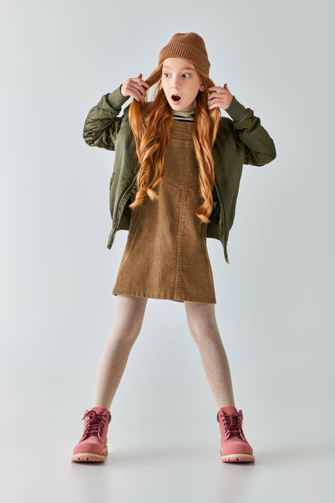 shocked preteen girl with long hair posing in stylish winter outfit and hat on grey backdrop - Photo, Image
