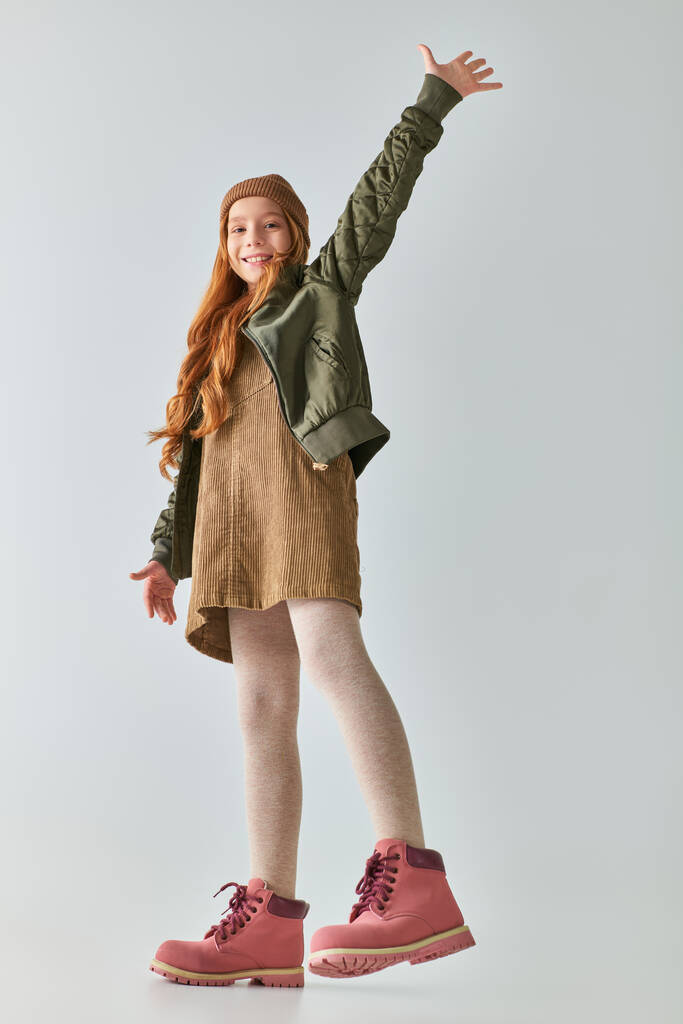χαρούμενο κορίτσι σε κομψό χειμωνιάτικο ντύσιμο με μπότες και καπέλο χαμογελώντας και κουνώντας το χέρι σε γκρι φόντο - Φωτογραφία, εικόνα