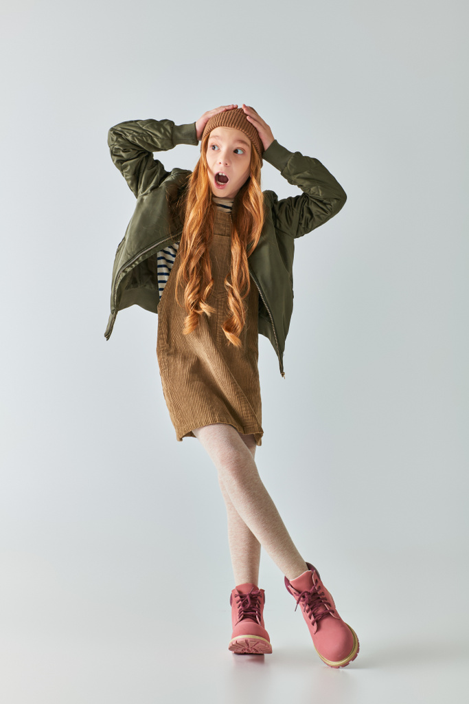 bang tiener meisje met lang haar in stijlvolle winter outfit en hoed schreeuwen op grijze achtergrond - Foto, afbeelding