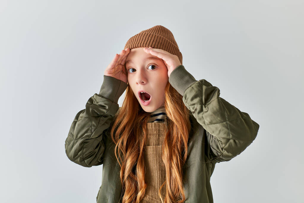 εκφραστική φοβισμένη κοπέλα σε κομψό χειμερινό ντύσιμο με πλεκτό καπέλο φουσκώνει μάγουλα σε γκρι φόντο - Φωτογραφία, εικόνα