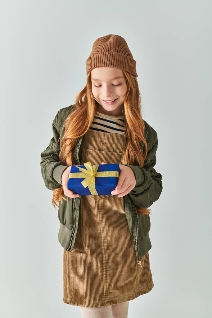 wesoła dziewczyna w stylowym stroju zimowym z kapeluszem gospodarstwa zawinięty prezent świąteczny na szarym tle - Zdjęcie, obraz