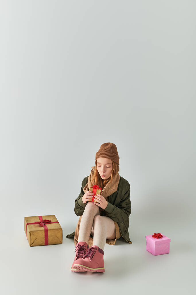 słodkie nastolatek dziewczyna w zimie strój z dzianiny kapelusz gospodarstwa Boże Narodzenie prezent i siedzi na szary - Zdjęcie, obraz