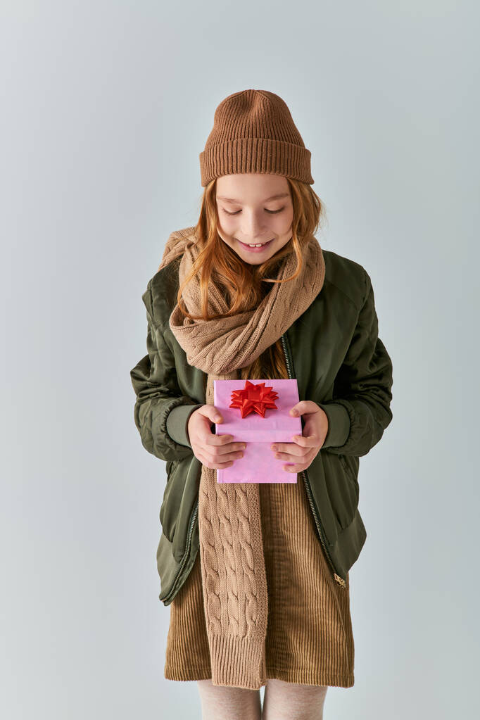happy preteen kid in stylish outfit with winter hat holding Christmas present and standing on grey - Photo, Image