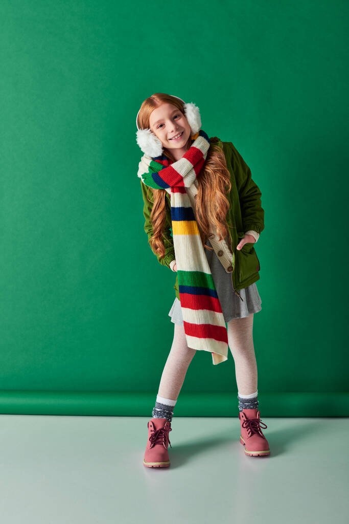 full length, χαρούμενο κορίτσι με ωτοασπίδες, ριγέ φουλάρι και χειμωνιάτικο ντύσιμο σε τυρκουάζ φόντο - Φωτογραφία, εικόνα
