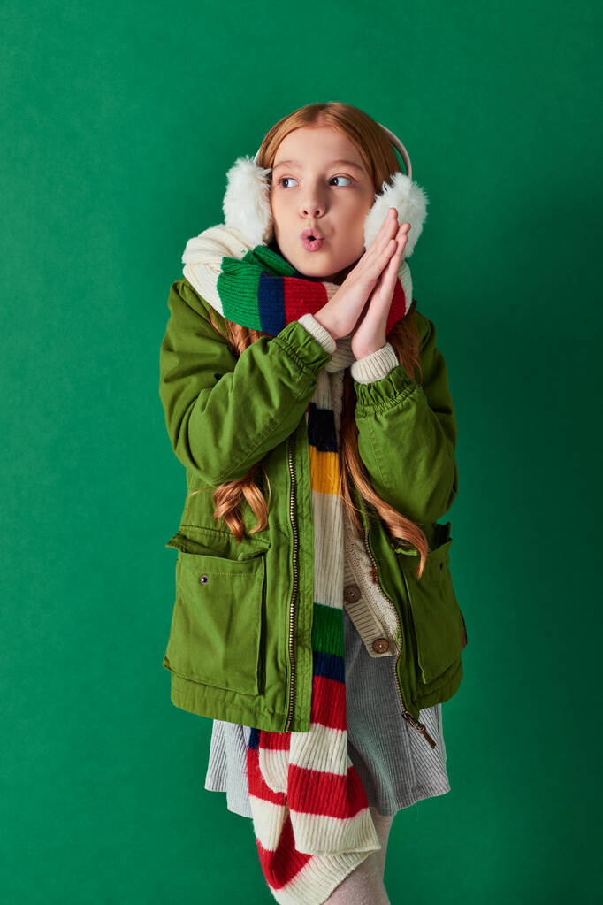 niña preadolescente en orejeras, bufanda rayada y ropa de invierno sensación de frío sobre fondo turquesa - Foto, imagen