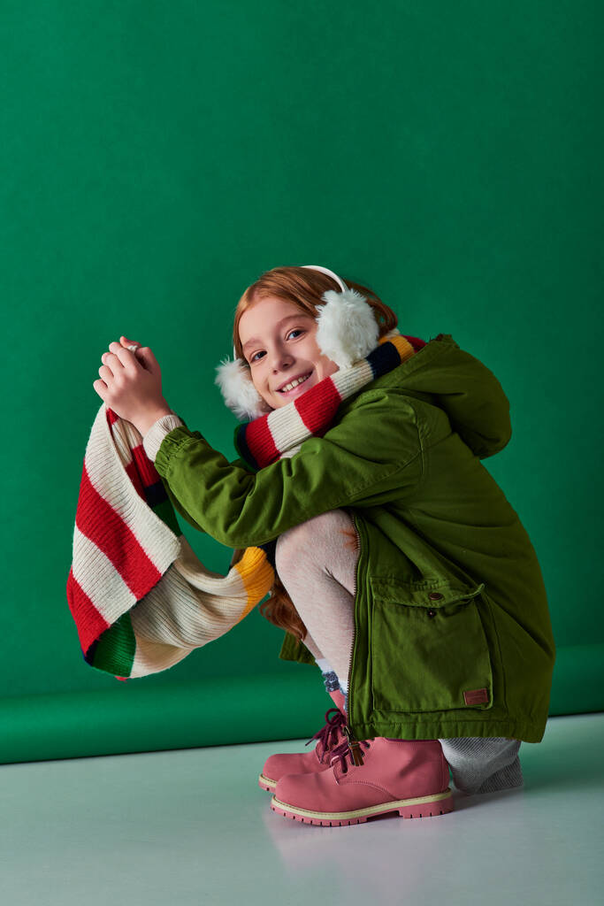 wesoły nastolatek dziewczyna w nauszniki, szalik w paski i strój zimowy siedzi na turkusowym tle - Zdjęcie, obraz