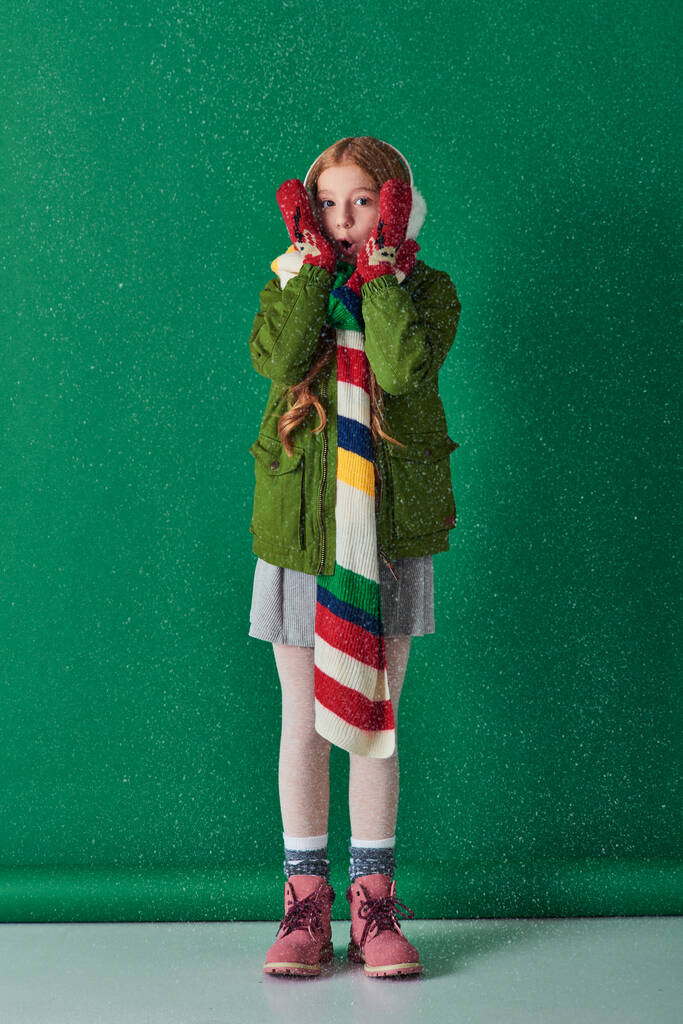 chica sorprendida en orejeras, bufanda y atuendo de invierno de pie bajo la nieve que cae en turquesa - Foto, imagen