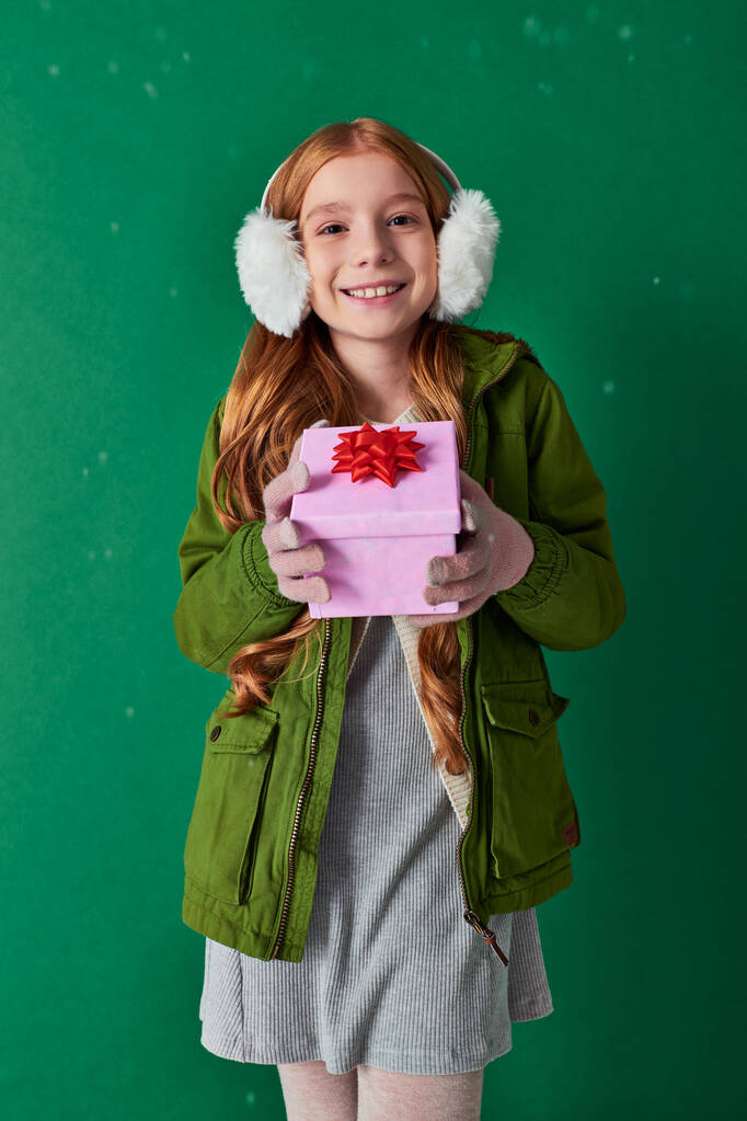 χαρούμενο κορίτσι σε ωτοασπίδες, κασκόλ και χειμωνιάτικη ενδυμασία κρατώντας Χριστουγεννιάτικο δώρο κάτω από το χιόνι - Φωτογραφία, εικόνα