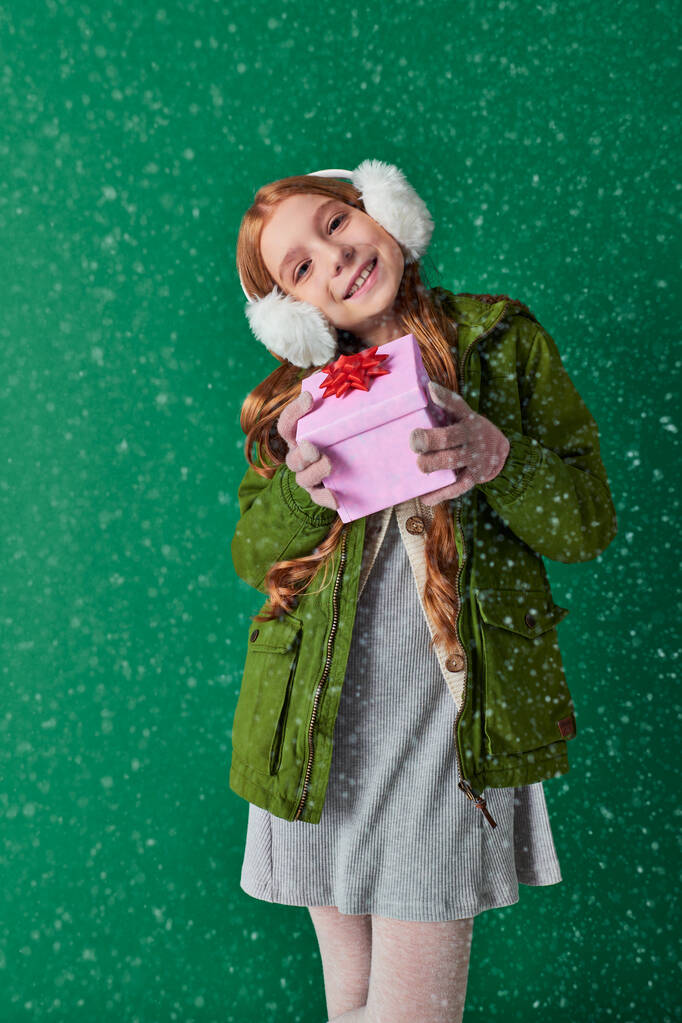 potěšená dívka v ušních manžetách, šála a zimní oblečení drží vánoční dárek pod padajícím sněhem - Fotografie, Obrázek