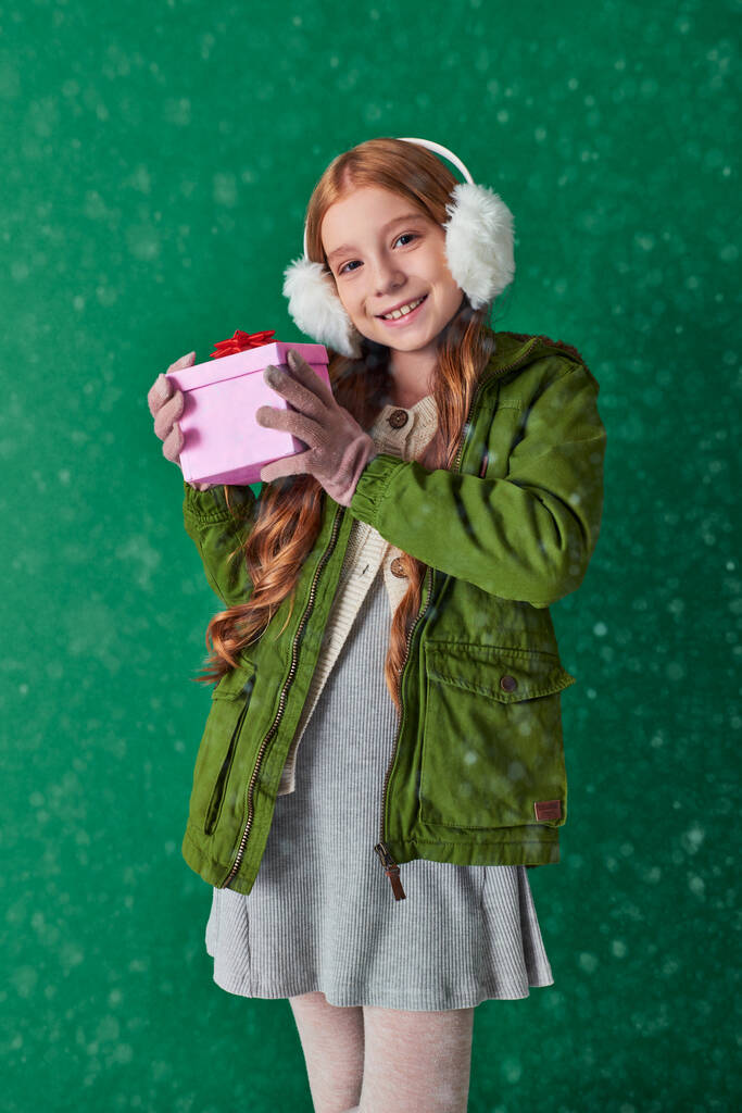 preteen ragazza in paraorecchie, sciarpa e abbigliamento invernale tenendo regalo di Natale sotto la neve caduta - Foto, immagini