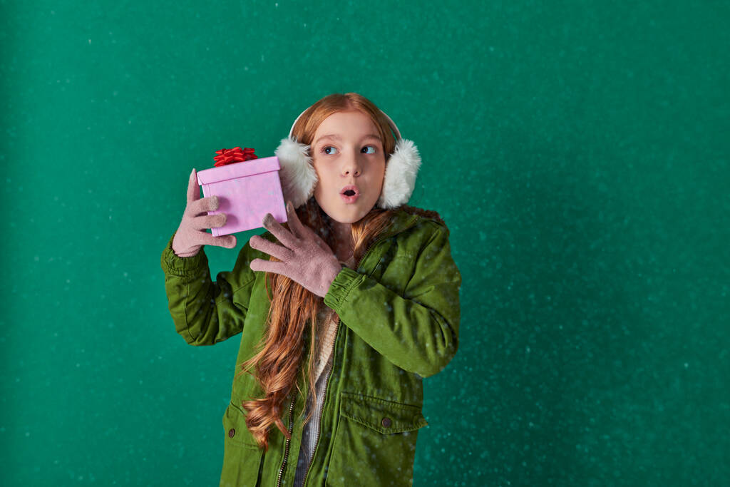 Любопытная девушка в наушниках, шарфе и зимнем наряде держит рождественский подарок под падающим снегом - Фото, изображение