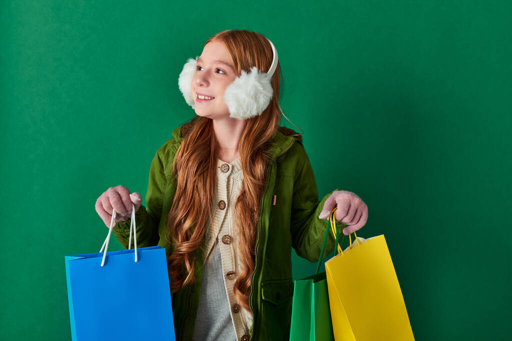 holiday season, happy girl in winter outfit and ear muffs holding shopping bags on turquoise - Photo, Image
