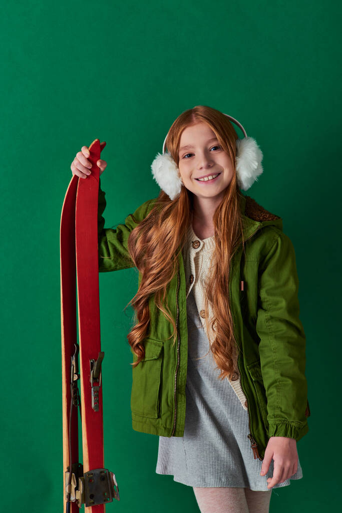 heureuse fille preteen dans des cache-oreilles et tenue d'hiver souriant et tenant des skis rouges sur fond turquoise - Photo, image